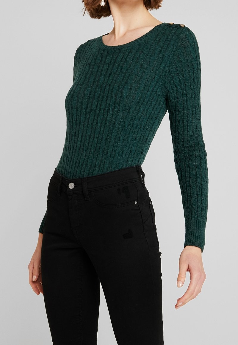 Femme portant un pull côtelé vert foncé manches longues avec boutons aux épaules et un pantalon noir taille haute sur un fond gris.