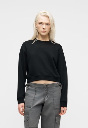 Ung kvinne med rett blondt hår som har på seg en svart croppet genser og grå cargobukser, står foran en ensfarget lys bakgrunn.