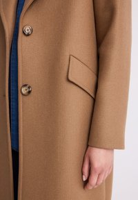 Beżowy wełniany płaszcz z klapowanymi kieszeniami i dwoma guzikami, noszony na dżinsowej koszuli, ukazujący prawą rękę i dłoń noszącego.