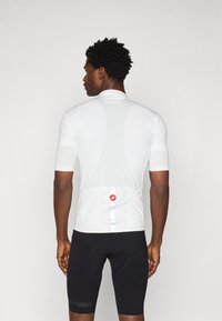 Maillot de cyclisme blanc à manches courtes avec un motif texturé, doté d'une poche arrière et d'un détail de logo rouge, associé à un cuissard de cyclisme noir.