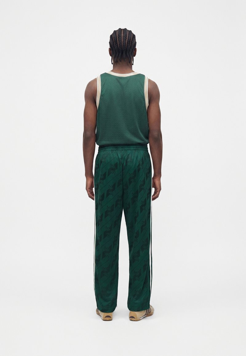 Mann mit geflochtenem Haar, der ein dunkelgrünes ärmelloses Trikot und passende gemusterte Hose trägt, steht mit dem Rücken zu einem weißen Hintergrund.