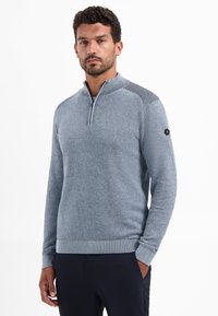 Man in een lichtblauwe sweater met rits tot net boven de borst en gestreepte schouderdetails, in donkere broek, staande voor een witte achtergrond.