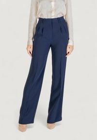 Pantalons plissés bleu marine à taille haute et à jambes larges, fabriqués dans un tissu lisse. Associés à une chemise à boutons de couleur claire.