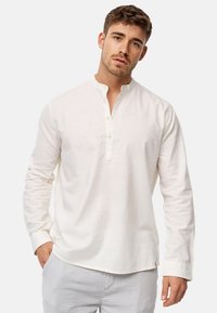 Long-sleeve, white linen shirt with a band collar and button placket. Soft texture with a relaxed fit and rounded hem detailing.