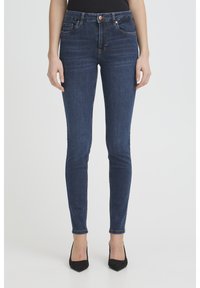 Sieviete tērpusies tumši zilās cieši pieguļošās džinsās un melnās augstpapēžu kurpēs ar smailiem galiem, stāvot pret vienkāršu gaišu fonu.