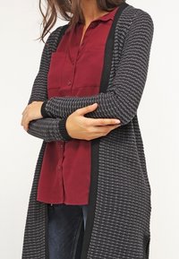 Long gray cardigan with a textured pattern, black trim, open front, and ribbed cuffs, layered over a burgundy button-up shirt.