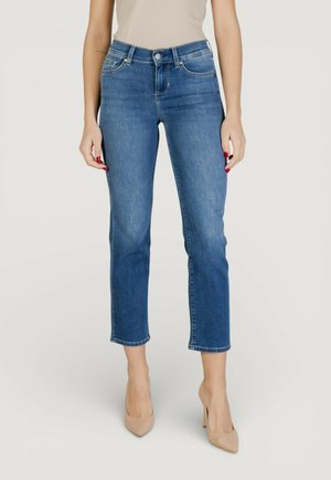 Femme portant un jean bleu court à jambes droites, un haut beige sans manches et des chaussures à talons hauts couleur nude, debout devant un fond uni.