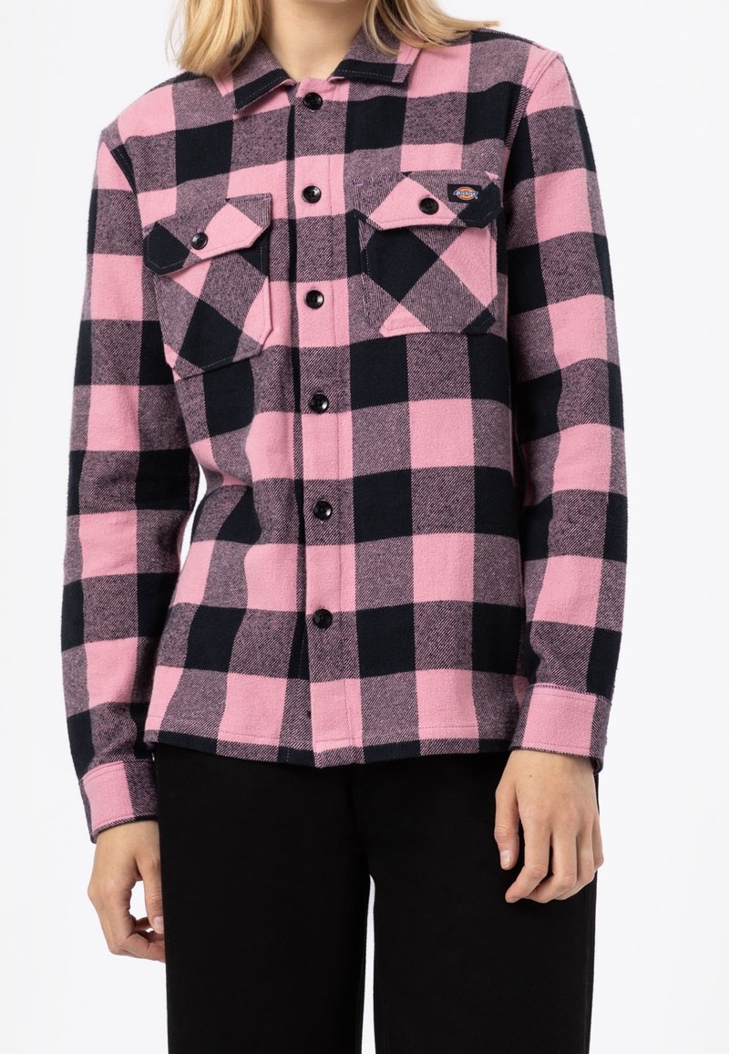 Camicia di flanella a quadrati rosa e neri con due taschini sul petto e bottoni neri. Il tessuto ha una consistenza morbida, con colletto e maniche lunghe.