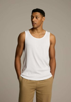 Biała bawełniana koszulka na ramiączkach z okrągłym dekoltem i szerokimi ramiączkami, w połączeniu z beżowymi spodniami. Gładki materiał i luźny krój.