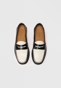Um par de mocassins de couro preto e branco com palmilhas castanhas, detalhes costurados e uma faixa preta na parte da frente sobre um fundo branco.