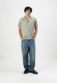 Kortsleiven lys grønn skjorte med bladmønster, kombinert med løse blå denimjeans og svarte sko, vist på en mannemodell.