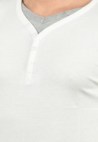 Valkoinen henley-paita, jossa on kolmesta napista muodostuva kaulus, teksturoidusta kankaasta ja kontrastivärinen harmaa kaulus, joka esittelee minimalistista muotoilua.
