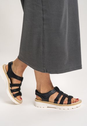 Black leather sandals featuring braided straps, adjustable fasteners, and a white textured sole, paired with a gray ankle-length skirt.