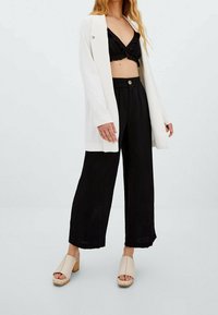 Femme portant un blazer blanc, un haut court noir, un pantalon large noir et des sandales à talons beiges, debout sur un fond clair uni.