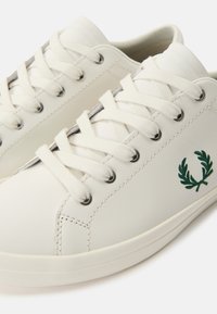Λευκά χαμηλά sneakers με λευκά κορδόνια, μεταλλικά ρηχές, και πράσινο κεντημένο λογότυπο στεφάνι δάφνης στη πλευρά.