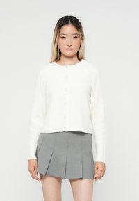 Vit kort cardigan gjord av mjukt, fluffigt tyg. Har rund halsringning, långa ärmar och sex pärlliknande knappar framtill.