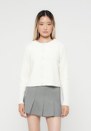 Hvit kort cardigan laget av myk, fluffy stoff. Har rund hals, lange ermer og seks perle-lignende knapper foran.