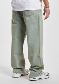 Jeans baggy de color verde claro con una cintura alta, que presentan bolsillos traseros y un desvanecimiento sutil, combinados con zapatillas deportivas blancas y verdes.