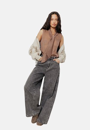 Femme aux longs cheveux foncés portant un haut sans manches à motifs, une veste légère drapée sur les épaules, un jean gris à jambes larges et des mocassins marron.