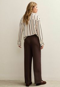 Gestreepte blouse met bruine en crèmekleurige verticale strepen, gecombineerd met losse bruine broeken. Het materiaal lijkt glad; de blouse heeft een knoopmanchet.