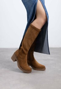 Botas de ante marrón hasta la rodilla con un tacón ancho, que presentan detalles de costuras y un acabado suave, combinadas con una falda de mezclilla con abertura.