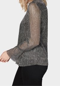 Langärmliges oberteil in Schwarz und silberner Metallic-Optik mit transparenten, strukturierten Ärmeln, getragen mit schwarzen Hosen, seitliche Ansicht.