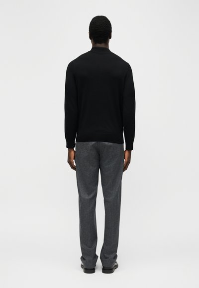 Άνδρας που στέκεται με την πλάτη, φοράει μαύρο πουκάμισο με μακριά μανίκια, γκρι παντελόνι και μαύρα παπούτσια, με φόντο λείο και ανοιχτό.