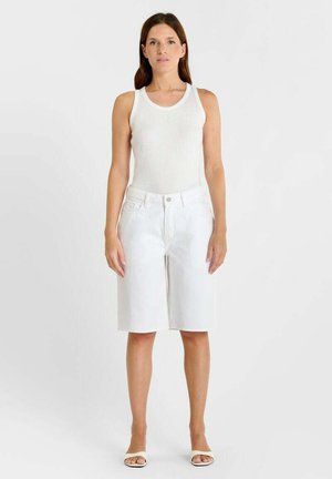 Frau steht aufrecht, trägt ein weißes ärmelloses Oberteil, weiße knielange Shorts und weiße Sandalen mit offenen Zehen vor schlichtem Hintergrund.