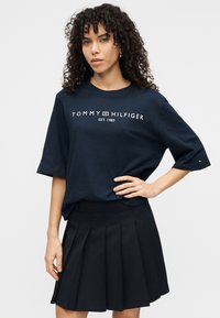 Μπλε μπλουζάκι από βαμβάκι με κοντά μανίκια, με λευκό λογότυπο "TOMMY HILFIGER EST. 1985", σε συνδυασμό με μαύρη πλισέ φούστα.