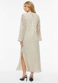 Lang, kremfarget kjole med svarte polkadotter, gjennomsiktig stoff, lange ermer, høy splitt og nøkkelholldetalj på ryggen. Fremhevet med svart kant.