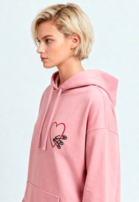 Felpa con cappuccio rosa realizzata in tessuto morbido, con un design di cuore rosso e cuciture nere, dotata di tasca a canguro e vestibilità rilassata.