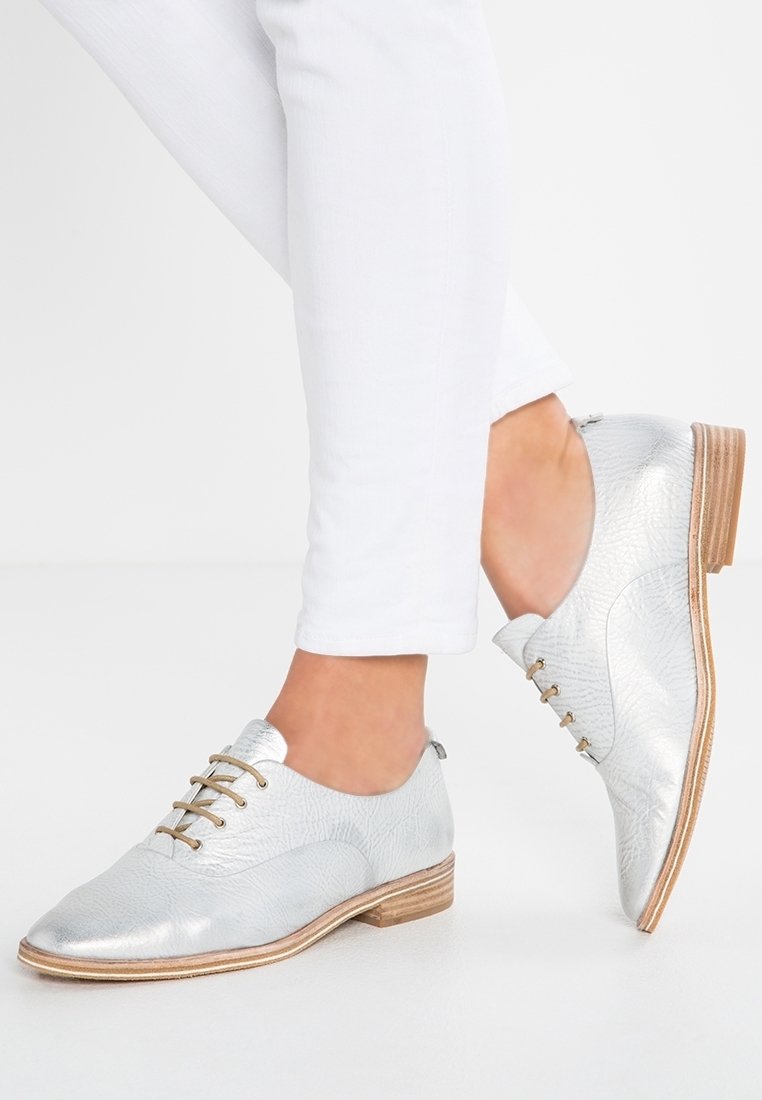 Chaussures Oxford en cuir argenté à lacets portées avec un pantalon blanc ajusté, montrant les deux pieds posés sur une surface blanche.