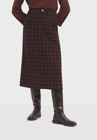 Geruite midi-rok in donkerrood en marineblauw, gemaakt van gestructureerde stof. Heeft een tailleband met een knoop en zijzakken. Draagt bijpassende bruine kniehoge laarzen.