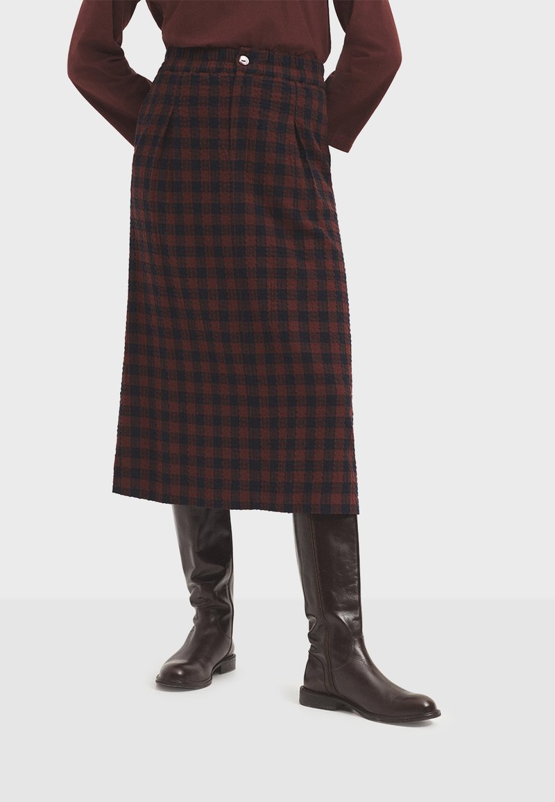 Geruite midi-rok in donkerrood en marineblauw, gemaakt van gestructureerde stof. Heeft een tailleband met een knoop en zijzakken. Draagt bijpassende bruine kniehoge laarzen.