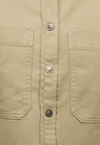 Μπεζ ύφασμα με δύο μπροστινές τσέπες και τρία μεταλλικά κουμπώματα με σκράτς σε κατακόρυφη ευθυγράμμιση στο κέντρο.