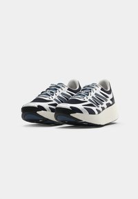 Et par hvide og marineblå atletiske sneakers med tykke såler og snørebånd, præsenteret på en ensfarvet lysegrå baggrund.