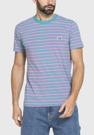 Uomo che indossa una maglietta lilla a maniche corte con strisce orizzontali turchesi, bianche e rosa e jeans di denim su uno sfondo semplice.