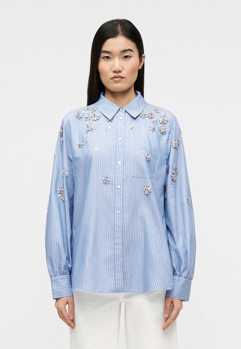 Γυναίκα που φοράει μπλε και άσπρο ριγέ πουκάμισο με ασημένιες ανθικές εφαρμογές, συνδυασμένο με άσπρο παντελόνι, όρθια μπροστά σε ένα απλό φόντο.