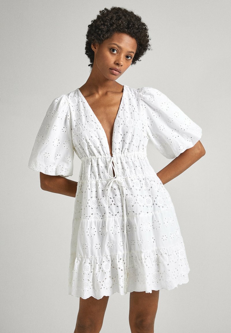 Robe en coton blanc avec des manches bouffantes, un décolleté en V profond et une taille à cordon. Présente des motifs en œillets et un ourlet évasé à étages.