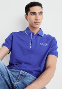 Fialová polokošeľa s golierom, s bielym pruhom a logom "Red Bull Racing". Nosená s svetlomodrými denimovými džínami.