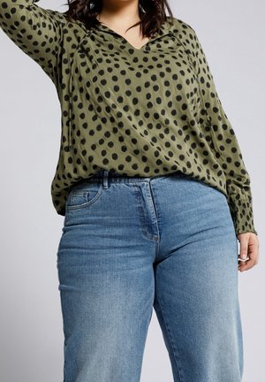 Personne grande taille portant un chemisier vert olive à pois noirs et un jean bleu en denim, debout devant un fond clair uni.