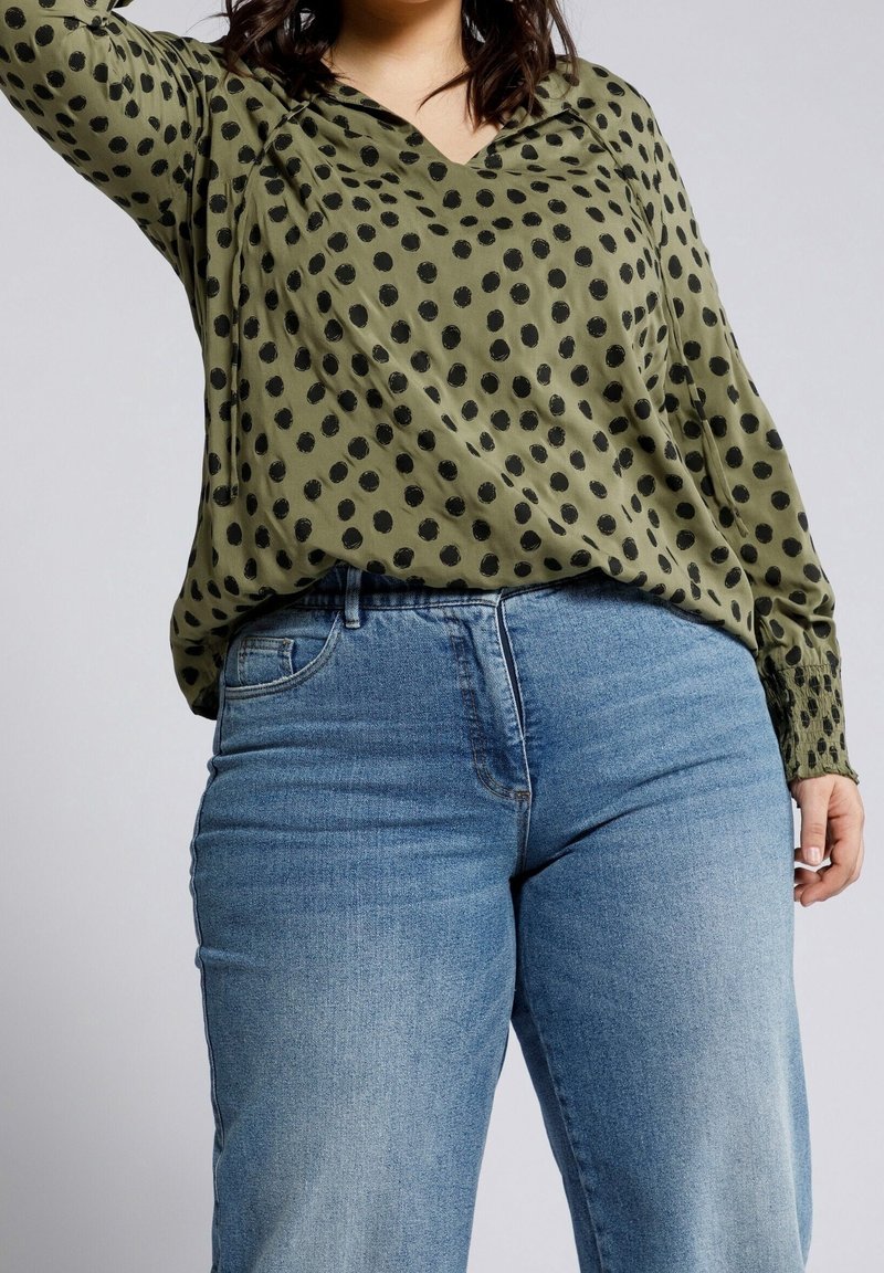 Personne grande taille portant un chemisier vert olive à pois noirs et un jean bleu en denim, debout devant un fond clair uni.