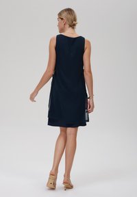 Marineblaues ärmelloses Kleid mit transparenter Überlage, glattem Stoff, knielang, seitlichen Schlitzen und minimalistischem Design. Getragen mit beigen Absatzschuhen.