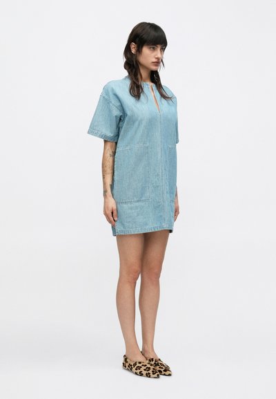 Γυναίκα που φορεί ανοιχτό μπλε ντένιμ φόρεμα με κοντά μανίκια, με μπροστινές τσέπες και μυτερές μπαλαρίνες με λεοπάρ σχέδιο, στέκεται μπροστά σε λευκό απλό φόντο.
