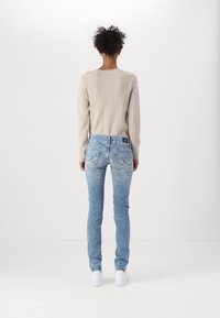 Pull en ribbed beige et jeans bleu clair avec poches arrière. Les jeans sont ajustés avec une légère décoloration ; des baskets blanches complètent la tenue.