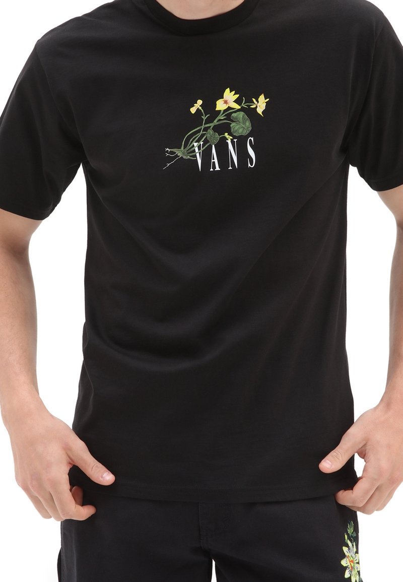 Personne portant un t-shirt noir Vans avec un graphisme de fleur jaune et le texte blanc Vans sur la poitrine, et un short noir avec broderie florale.