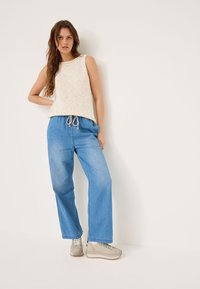 Top in maglia senza maniche di un beige chiaro con scollatura rotonda, abbinato a pantaloni in denim azzurri larghi e sneakers grigie con suola testurizzata.