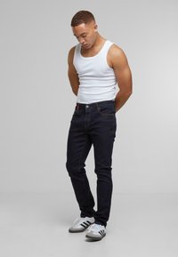 Sötét farmernadrág, slim fit szabással, kontraszt varrásokkal és fém gombbal. Fehér ujj nélküli felsővel és szürke-fehér sportcipővel párosítva.