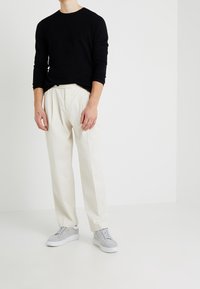 Camicia nera a maniche lunghe con texture, pantaloni plissettati color crema e scarpe da ginnastica grigie con lacci blu. Uno sfondo neutro mette in risalto l'outfit.