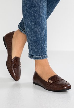 Osoba nosząca ciemnoniebieskie dżinsy z denimu oraz brązowe, płaskie loafersy o strukturze krokodyla z drobnymi metalowymi guzikami.